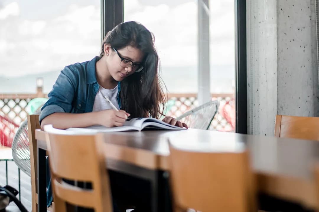 Estudante de óculos escrevendo em um livro aberto, sentada à mesa